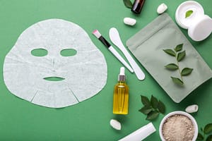 Fabric cosmetic face mask with set of cosmetics clay mask serum oil spatula brush, moisturizing cream in jar. Beauty spa treatments for facial skin care, cosmetology on green background with leaves